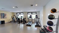 The fitness center at Copperwood at Princeton is a well-appointed exercise facility designed to support a healthy and active lifestyle. The space features a clean, modern aesthetic with crisp white walls, recessed lighting, and a durable, easy-to-maintain floor surface suited for high-traffic use. Natural light floods the room through multiple large windows framed in dark trim, offering pleasant views of the surrounding greenery and creating an energizing atmosphere for workouts. The cardio area is equipped with treadmills, elliptical machines, and a stationary bike, several of which feature integrated display screens for entertainment or performance tracking. A rack of free weights lines one side of the room, providing options for strength and resistance training. The ceiling-mounted HVAC system ensures consistent airflow and comfort throughout the space. The layout is thoughtfully organized to allow for ease of movement between equipment, making the facility accessible and functional for residents of varying fitness levels. Overall, the fitness center at Copperwood at Princeton delivers a practical and inviting environment for daily exercise.