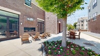 Located at 308 E Second St in Hummelstown, PA, this outdoor courtyard is a thoughtfully designed communal space nestled between brick-clad buildings. The courtyard offers a variety of distinct activity zones, making it a versatile gathering spot for relaxation and socializing. A built-in fireplace set against the brick exterior wall serves as a natural focal point, surrounded by a cluster of classic Adirondack chairs ideal for lounging. Adjacent landscaped beds filled with colorful flowering plants and a mature shade tree add natural beauty and a sense of tranquility to the space. String lights are strung overhead throughout the courtyard, creating a warm and inviting ambiance that extends into the evening hours. Multiple wooden dining tables and chairs, along with a picnic table, provide ample seating for outdoor meals and gatherings. A gas grill station supports outdoor cooking activities, while a dedicated cornhole area offers casual recreation. Bike racks conveniently located along the perimeter provide secure storage for residents. The courtyard is framed by the surrounding multi-story residential buildings, offering a sense of enclosure while remaining open to the sky, allowing abundant natural light throughout the day.