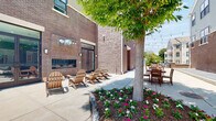 Located at 308 E Second St in Hummelstown, PA, this outdoor courtyard is a thoughtfully designed communal space nestled between brick-clad buildings. The courtyard offers a variety of distinct activity zones, making it a versatile gathering spot for relaxation and socializing. A built-in fireplace set against the brick exterior wall serves as a natural focal point, surrounded by a cluster of classic Adirondack chairs ideal for lounging. Adjacent landscaped beds filled with colorful flowering plants and a mature shade tree add natural beauty and a sense of tranquility to the space. String lights are strung overhead throughout the courtyard, creating a warm and inviting ambiance that extends into the evening hours. Multiple wooden dining tables and chairs, along with a picnic table, provide ample seating for outdoor meals and gatherings. A gas grill station supports outdoor cooking activities, while a dedicated cornhole area offers casual recreation. Bike racks conveniently located along the perimeter provide secure storage for residents. The courtyard is framed by the surrounding multi-story residential buildings, offering a sense of enclosure while remaining open to the sky, allowing abundant natural light throughout the day.