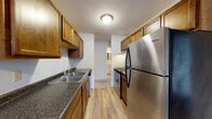 This single-floor unit offers a straightforward, functional layout revealed clearly in its 3D floor plan. The galley-style kitchen is well-equipped, featuring warm medium-toned wood cabinetry both above and below the counters, dark granite-style countertops, a double stainless steel sink, and stainless steel appliances including a refrigerator and an electric range with overhead ventilation. Light wood-look flooring runs throughout the kitchen, lending a clean, modern contrast to the cabinetry. The kitchen connects directly to adjacent living areas, creating an open flow through the unit.

The living area is generously proportioned, finished with neutral carpet and bright white walls, accented by dark wood baseboards. A large sliding glass door or wide window draws in natural light and provides outdoor views, while a wall-mounted AC unit and baseboard heating ensure year-round climate control. A textured ceiling adds dimension overhead.

The bedrooms are similarly finished with plush carpet, ceiling fans with integrated lighting, individual windows with blinds, and baseboard heating. The bathroom features full white tile surrounding a tub-shower combination, a toilet, and a vanity, maintaining a clean and classic look throughout. Overall, the unit presents a practical and cohesive living environment.