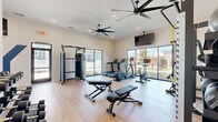 The Yardly Town Center fitness facility at 151 S Buddy Hayes Blvd in Anna, TX, offers a thoughtfully designed, single-floor workout space that balances functionality with a clean, modern aesthetic. The gym features light hardwood-style flooring throughout, crisp white walls, and recessed lighting complemented by large ceiling fans that ensure consistent airflow and comfort during workouts.

Large floor-to-ceiling windows and sliding glass doors line the perimeter, flooding the space with abundant natural light and providing views of the surrounding outdoor landscape. The open layout is well-organized, housing a comprehensive range of equipment to support diverse fitness routines. Cardio options include treadmills, a stationary bike, and an elliptical, while strength training is well represented with a cable machine, a power rack with weight plates, adjustable benches, and a full dumbbell rack. A dedicated storage shelf along one wall holds medicine balls, stability balls, and other accessories.

A wall-mounted flat-screen television provides entertainment or instructional content during workouts. The space also includes a water fountain for convenience. The "Yardly Town Center" branding is prominently displayed on the wall, reinforcing the community identity of this amenity. The overall ambiance is bright, inviting, and purposefully equipped to serve a wide range of fitness needs.