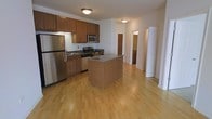 This single-floor residence is anchored by a well-appointed kitchen that serves as the focal point of the open-concept living space. The kitchen is outfitted with a full suite of stainless steel appliances, including a refrigerator, gas range, and built-in microwave, all complemented by rich wood cabinetry that extends to the upper walls for ample storage. Granite countertops add a polished finish, while a central kitchen island provides additional workspace and casual dining potential. The warm-toned hardwood flooring flows seamlessly throughout the main living area, lending continuity and a sense of spaciousness to the layout. Flush-mount ceiling light fixtures provide consistent overhead illumination across the space. A hallway visible from the kitchen leads to additional areas of the unit, suggesting a functional and well-organized floor plan. The overall ambiance is clean and neutral, with light-colored walls that enhance brightness and create a versatile backdrop suited to a variety of interior styles.