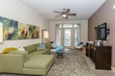 This single-story residence offers a well-designed layout that balances functionality and contemporary style across multiple rooms. The kitchen serves as a central highlight, featuring rich dark wood cabinetry extending to the upper walls, black appliances including a range, microwave, and dishwasher, and light-toned countertops. A kitchen island provides additional prep and dining space, while wood-look flooring and pendant lighting with track fixtures overhead add a polished finish. The living room is generously proportioned, with a ceiling fan, large windows and glass doors that invite natural light and offer outdoor views, and neutral carpet underfoot. A separate sitting area features warm brown accent walls and a wall-mounted television above a dark media console. Multiple bedrooms are present, each carpeted and equipped with ceiling fans. Bedrooms connect to walk-in closets, some of which include in-unit laundry. Bathrooms are finished with tile flooring, dark vanity cabinetry, light countertops, and a full tub with shower in one, while another features an elongated vanity with ample storage. Throughout the residence, a cohesive modern aesthetic is maintained through consistent use of dark wood tones and neutral wall colors.