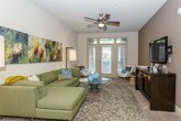 This single-story residence offers a well-designed layout that balances functionality and contemporary style across multiple rooms. The kitchen serves as a central highlight, featuring rich dark wood cabinetry extending to the upper walls, black appliances including a range, microwave, and dishwasher, and light-toned countertops. A kitchen island provides additional prep and dining space, while wood-look flooring and pendant lighting with track fixtures overhead add a polished finish. The living room is generously proportioned, with a ceiling fan, large windows and glass doors that invite natural light and offer outdoor views, and neutral carpet underfoot. A separate sitting area features warm brown accent walls and a wall-mounted television above a dark media console. Multiple bedrooms are present, each carpeted and equipped with ceiling fans. Bedrooms connect to walk-in closets, some of which include in-unit laundry. Bathrooms are finished with tile flooring, dark vanity cabinetry, light countertops, and a full tub with shower in one, while another features an elongated vanity with ample storage. Throughout the residence, a cohesive modern aesthetic is maintained through consistent use of dark wood tones and neutral wall colors.