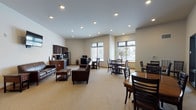 This single-floor community common area is thoughtfully designed to serve as a welcoming hub for socializing, dining, and relaxation. The open-plan layout is anchored by neutral-toned walls, light carpeting, and recessed lighting throughout, creating a bright and inviting atmosphere. Large windows allow natural light to filter in, offering views to the surrounding exterior.

The lounge area is furnished with rich brown leather sofas, a patterned accent chair, and wooden side tables, all arranged around a wall-mounted flat-screen television — ideal for casual gatherings. Adjacent to the lounge is a functional kitchenette equipped with dark wood cabinetry, a double stainless steel sink, a full-size stainless steel refrigerator, and a coffee station, providing convenient amenities for residents or guests.

The dining area features multiple solid wood tables with ladder-back chairs, accommodating both small and larger groups. Large glass panel windows and a sliding glass door look directly into an adjacent fitness room, maintaining a sense of openness and connectivity between spaces. A separate dining nook with a potted indoor tree adds a natural, calming touch near a bright exterior-facing window. The overall design balances practicality with comfort, making this a versatile and functional communal space.