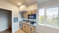 This single-floor unit offers a thoughtfully designed layout with contemporary finishes throughout. The kitchenette serves as a functional centerpiece, featuring warm wood-tone cabinetry with matte black hardware, a grey quartz-style countertop, an undermount sink with a black faucet, a built-in microwave, an induction cooktop, and a compact refrigerator below the counter — providing efficient use of space without sacrificing style. An electrical panel is also conveniently recessed into the kitchen wall. The main living area is bathed in natural light, thanks to large windows that frame views of the surrounding neighborhood and greenery. Light hardwood-style flooring runs continuously through the space, lending a clean, airy aesthetic that complements the neutral grey walls and white trim. A hallway connects the living area to additional rooms, with multiple doorways providing clear separation between spaces. The bathroom features a walk-in shower with white surround walls, a matte black showerhead and valve, and coordinating black accent hooks, maintaining the consistent modern design language found throughout the unit. Recessed lighting and HVAC venting are neatly integrated into the ceilings and walls, contributing to an overall polished and move-in-ready feel.