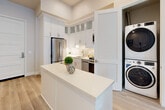 This thoughtfully designed one-bedroom, one-bathroom residence offers a bright and contemporary living experience across a single floor. The open-concept layout seamlessly connects the kitchen, dining area, and living room, creating a fluid and functional space ideal for everyday living and entertaining.

The kitchen is a standout feature, outfitted with full stainless steel appliances including a gas range, built-in microwave, dishwasher, and refrigerator. Crisp white shaker cabinetry extends to the ceiling with glass-front upper panels, complemented by a subway tile backsplash and a white quartz countertop island. A concealed stacked washer and dryer unit adds everyday convenience. Light hardwood flooring runs throughout, tying the spaces together cohesively.

The dining area provides a dedicated space between the kitchen and living room, while the living room impresses with soaring ceilings and floor-to-ceiling windows that flood the space with natural light and frame open city and neighborhood views.

The bedroom is a serene retreat featuring tall windows with outdoor views and a walk-in closet with built-in shelving. The bathroom is finished with white cabinetry, a quartz vanity, an illuminated mirror, and matching light flooring, maintaining the clean, modern aesthetic found throughout the residence.