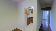 This single-floor residence offers a straightforward and functional layout designed for comfortable everyday living. The space is characterized by clean, neutral wall tones throughout, creating a bright and open atmosphere that makes each area feel welcoming.

The kitchen is accessible through a defined doorway, providing a sense of separation from the main living areas. Inside, warm honey-toned wood cabinetry lines the walls, offering ample storage space above and below the countertops. The dark countertop provides a subtle contrast to the cabinetry, while the light-tiled flooring keeps the kitchen feeling bright and easy to maintain. A ceiling-mounted flush light fixture ensures the kitchen is well-lit.

The hallway connecting the rooms features rich, dark hardwood flooring that adds warmth and character to the overall aesthetic. Classic white baseboards trim the walls throughout, lending a clean, finished look to each space. The corridor leads to additional rooms, hinting at a practical and well-organized floor plan.

Overall, this residence presents a functional and tidy living environment, with consistent design details — from the wood cabinetry to the hardwood floors — that tie the space together cohesively.