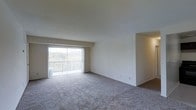 This single-floor space features an open-concept living area designed to maximize functionality and comfort. The room is finished with wall-to-wall carpeting in a neutral tone, providing a soft and cohesive foundation throughout. Smooth, white walls offer a clean and understated backdrop, making the space highly adaptable for a variety of interior styles.

A standout feature of the living area is the full-width sliding glass door fitted with vertical blinds, which opens directly onto a private balcony. This connection to the outdoors allows for generous natural light to fill the interior while offering views of surrounding greenery, creating a pleasant and airy atmosphere. The balcony itself provides an outdoor retreat accessible directly from the main living space.

An adjacent alcove or secondary area is partially visible, suggesting additional functional space that could serve as a dining area or secondary room. The layout flows openly, lending a sense of spaciousness to the overall footprint. The unit's simple, well-maintained finishes and practical design make it a comfortable and move-in ready living environment.