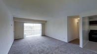 This single-floor space features an open-concept living area designed to maximize functionality and comfort. The room is finished with wall-to-wall carpeting in a neutral tone, providing a soft and cohesive foundation throughout. Smooth, white walls offer a clean and understated backdrop, making the space highly adaptable for a variety of interior styles.

A standout feature of the living area is the full-width sliding glass door fitted with vertical blinds, which opens directly onto a private balcony. This connection to the outdoors allows for generous natural light to fill the interior while offering views of surrounding greenery, creating a pleasant and airy atmosphere. The balcony itself provides an outdoor retreat accessible directly from the main living space.

An adjacent alcove or secondary area is partially visible, suggesting additional functional space that could serve as a dining area or secondary room. The layout flows openly, lending a sense of spaciousness to the overall footprint. The unit's simple, well-maintained finishes and practical design make it a comfortable and move-in ready living environment.