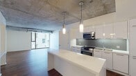 Located on the iconic Sunset Strip in West Hollywood, this single-floor unit showcases a striking industrial-modern aesthetic defined by exposed concrete ceilings throughout, rich dark hardwood flooring, and crisp white walls that create a bold visual contrast. The open-concept kitchen is a standout feature, outfitted with high-gloss white cabinetry, a grey tile backsplash, stainless steel appliances including a range and built-in microwave, and a generous white waterfall island that anchors the space. Cylindrical pendant lights hang from the raw concrete ceiling, adding a designer touch. The living area benefits from expansive floor-to-ceiling windows that flood the space with natural light and frame views of the surrounding West Hollywood cityscape. The bedroom continues the industrial-chic theme with the same concrete ceiling and large windows with roller shades for privacy. A built-in bench near the entryway adds functional detail. The bathrooms are finished in a clean, contemporary style featuring tiled tub-shower combinations with glass enclosures, undermount sinks, dark wood vanities with white countertops, and frameless mirrors, delivering a polished, modern feel throughout.