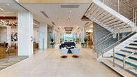 This expansive commercial space presents a sophisticated, contemporary design defined by an open, flowing layout and an abundance of natural light. The main lobby corridor stretches impressively from the glass-paneled entrance to floor-to-ceiling curtain walls at the rear, framing outdoor views of landscaped grounds and a pool area beyond. Large-format polished porcelain tile flooring runs throughout, amplifying the sense of scale and brightness.

Soaring ceilings with recessed linear LED lighting and coffered wood-accented details add warmth and architectural depth to the otherwise crisp white interior. A dramatic stacked-stone accent column anchors the space, complementing a sleek floating staircase with steel cable railings. Multiple lounge seating areas are arranged throughout, furnished with contemporary sofas and accent chairs in neutral and navy tones.

Glass-walled rooms line the perimeter, including a well-appointed business center featuring built-in wood shelving, a row of workstations with ergonomic seating, and a dedicated lounge nook. Potted tropical plants positioned throughout add organic texture and vitality. The overall ambiance is polished, professional, and welcoming, combining modern commercial design with resort-inspired elegance.