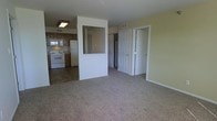 This single-floor unit offers an open layout that visually connects the living area to the kitchen. The living space is finished with wall-to-wall carpet in a neutral tone, creating a comfortable and cohesive feel throughout. Pale yellow walls provide a warm, understated backdrop that enhances the sense of brightness in the space.

The kitchen is situated at the rear and features dark wood cabinetry, a white electric range, a refrigerator, and a built-in microwave, all set atop tile flooring that contrasts with the carpeted living area. A pass-through window or open partition wall connects the kitchen visually to the main living space, improving sightlines and airflow between the two areas.

A central structural column and partial wall divide the living area from a hallway, where multiple white six-panel doors provide access to additional rooms, including what appears to be a closet and at least one other room. Ceiling-mounted lighting and overhead HVAC vents indicate climate control is integrated throughout. The overall layout is practical and efficiently organized for everyday living.