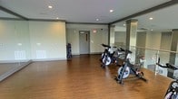 This single-floor fitness facility is designed to support an active lifestyle with a clean, functional layout. The space features warm hardwood-style flooring that extends throughout, providing a durable and attractive surface suited for a variety of exercise activities. Recessed lighting in the ceiling ensures the area is well-lit, creating a bright and energizing environment for workouts.

The cycling area is a standout feature, housing multiple spinning bikes arranged to accommodate group or individual training sessions. A rack nearby offers organized storage for accessories. Structural columns with contrasting trim details add an architectural element to the space, while large mirrored panels along one wall visually expand the room and allow users to monitor their form during exercise.

The facility is finished with neutral gray walls that contribute to a modern, professional atmosphere. A secured access door provides a dedicated entry and exit point, and a staircase visible along one side suggests convenient connectivity within the broader building. Overall, the space strikes a balance between practicality and comfort, offering a streamlined fitness environment that is both inviting and purpose-built for exercise.