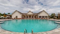 Located at 101 Tomshire Dr in Johns Island, SC, this single-story community clubhouse showcases a charming farmhouse-inspired architectural design characterized by crisp white board-and-batten siding, a standing-seam metal roof with multiple gabled peaks, and warm brick accents along the base of the building. The centerpiece of the facility is a generously sized outdoor swimming pool with clear turquoise water, accessible via stainless steel entry rails and surrounded by an expansive stone-tile pool deck. The deck offers ample space for relaxation, furnished with sling-style lounge chairs positioned to take in views of the surrounding residential community. A covered porch area extends from the main structure, providing a shaded retreat equipped with comfortable wicker seating, including a sectional sofa, armchair, and ottoman with neutral cushions and colorful accent pillows. Large French doors with grid-pane windows connect the interior to the outdoor spaces, flooding both areas with natural light and offering direct visual access to the pool. The broad, light-toned stone pavers throughout the patio and covered areas lend a polished, cohesive look to the outdoor environment. The overall ambiance is relaxed yet refined, reflecting the welcoming character of the Johns Island community.