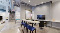 This stylish community amenity space is thoughtfully designed to offer a variety of social and recreational experiences within a single, open floor plan. The layout flows seamlessly between distinct zones, beginning with a welcoming lounge area anchored by a curved sectional sofa, rounded ottomans, and bold yellow-and-grey geometric patterned carpet. Soaring floor-to-ceiling windows flood this area with abundant natural light and provide views of the surrounding streetscape, while an eye-catching circular teal ceiling detail adds a sculptural architectural element overhead.

Adjacent to the lounge, a full-size pool table with a vibrant turquoise felt surface sits beneath a modern circular pendant light, with a wall-mounted cue rack finished in striking yellow providing a pop of color. Exposed ductwork and polished concrete flooring lend an industrial character that contrasts elegantly with the refined furnishings throughout.

A dedicated media and bar-height seating area features blue upholstered counter stools at a sleek white table, a large wall-mounted flat-screen TV, and textured accent wall panels. A semi-private meeting alcove off to the side offers a bright workspace bathed in natural light from large windows, complete with a conference table, blue chairs, and a branded glass partition wall. Together, these zones create a dynamic and functional community gathering space.