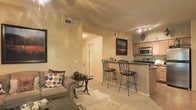 This single-floor space showcases an open-concept layout that connects the living area and kitchen, creating a cohesive and functional environment. The living area features warm beige carpeting, a comfortable sofa accented with decorative pillows, and a small accent table, all complemented by framed wall art that adds a touch of character. Recessed lighting and track lighting illuminate the space effectively, enhancing the warm, neutral color palette of the walls and ceilings.

The kitchen is positioned adjacent to the living area and is separated by a breakfast bar fitted with a dark countertop and accommodating two wrought-iron bar stools. The kitchen includes wood-toned cabinetry, a stainless steel microwave, and a range, providing practical cooking amenities. The countertops offer ample prep space, and the layout promotes easy interaction between the kitchen and living areas.

A hallway visible from the main living area leads to additional rooms, suggesting a well-organized floor plan. The overall ambiance is warm and inviting, with consistent neutral tones, track and recessed lighting, and an efficient use of the available space throughout.