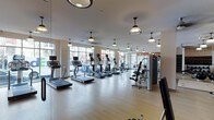 This fitness center offers a thoughtfully designed workout environment that balances functionality with an inviting aesthetic. The space is laid out on a single level, providing an open and unobstructed floor plan that accommodates a comprehensive range of exercise equipment. Rows of treadmills and elliptical machines are positioned along the perimeter, each unit equipped with individual screens for entertainment during cardio sessions. Strength training machines are organized toward the rear of the space, offering users a variety of resistance and weight options.

Large floor-to-ceiling windows line one wall, flooding the interior with generous natural light and offering views of the surrounding streetscape, creating an energizing atmosphere. The warm-toned hardwood-style flooring adds a clean, polished look underfoot while providing durability suited to high-traffic use. Overhead, a series of classic dome-shaped pendant lights are evenly distributed across the ceiling, complementing the bright, airy feel of the room. Ceiling fans are also integrated into the layout, promoting air circulation throughout the space. Full-length mirrors along one wall enhance the sense of openness and allow users to monitor their form during workouts.
