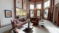 This expansive single-floor space showcases an impressive great room defined by dramatically tall ceilings supported by exposed wooden beams and a striking floor-to-ceiling stone fireplace with brick detailing. A ceiling fan and Western-themed artwork accent the soaring walls, while tufted leather sofas, cowhide accents, and wrought iron furnishings reinforce the space's rustic character. Multiple seating arrangements create distinct conversation areas centered around a wood-and-metal coffee table.

An adjoining dining area benefits from towering wraparound windows dressed in rich chocolate-brown drapes, flooding the space with natural light and offering views of the surrounding exterior. Tall potted palms flank the window walls, adding a tropical contrast to the Western aesthetic. A dark wood dining table with carved chairs sits beneath an ornate candelabra-style chandelier, complementing the nearby sitting area with neutral wingback chairs.

The kitchen opens to the main living area via a pass-through counter with granite countertops, wood cabinetry, and roman shades over wood-slatted blinds. The layout promotes easy interaction between the kitchen and gathering spaces, making this a highly functional and inviting communal environment overall.