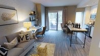 This single-floor space showcases a thoughtfully designed open-concept layout that seamlessly integrates the living, dining, and workspace areas. Dark hardwood-style flooring runs throughout, grounding the space with a warm, contemporary aesthetic. The living area features a neutral-toned sofa accented with decorative pillows and a distinctive gold geometric side table, adding a touch of glamour to the design.

A white brick accent wall serves as a focal point, adorned with floating black shelves displaying curated décor. Tucked against this wall is a compact desk area, making efficient use of the space for a functional home office nook. The dining area is anchored by a marble-topped table with a sleek industrial-style base, accompanied by elegant upholstered chairs.

Natural light floods the space through a large sliding glass door dressed with flowing patterned curtains, offering direct access to an outdoor patio furnished with seating. A side window with blinds provides additional brightness. The overall ambiance is modern and refined, balancing practicality with sophisticated style throughout.