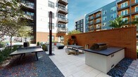 Located at 1701 Central Ave in the heart of Saint Petersburg, Florida, this thoughtfully designed outdoor courtyard serves as a vibrant communal gathering space. The area is organized into distinct functional zones, each offering its own character and purpose. A dedicated grilling station anchors one end of the space, outfitted with a built-in grill and an adjacent picnic-style dining table with matching benches, all set against a warm, textured brick accent wall. A wall-mounted screen adds an entertainment element to this outdoor dining area. Paved in large-format light stone tiles and complemented by gravel accents, the courtyard's hardscape creates a clean, contemporary aesthetic. Landscaping plays a central role throughout, with lush plantings including tropical grasses, bromeliads, palms, and ornamental shrubs lining the walkways and building perimeters. A cobblestone pathway leads through a garden corridor flanked by colorful foliage and bright orange accent seating, providing an inviting transition between zones. Slim modern light bollards punctuate the landscape. The courtyard is enclosed by the surrounding multi-story buildings, creating a sheltered, semi-private atmosphere while still remaining open to the Florida sky. The overall ambiance balances urban sophistication with natural warmth, making it a welcoming outdoor retreat.