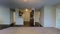 This single-floor space showcases an open-concept design that seamlessly connects the living area and kitchen. The living space features plush neutral carpeting, while the kitchen and adjoining areas transition to dark hardwood-style flooring, creating a distinct yet cohesive visual contrast. The kitchen is equipped with dark wood cabinetry, a white refrigerator, and a peninsula that doubles as a breakfast bar, providing both additional counter space and a casual dining option. A secondary kitchen counter with a sink adds further functionality to the layout. The textured ceiling adds a subtle architectural detail throughout the space, and recessed and flush-mount lighting ensures the interiors are well-lit. Neutral gray walls contribute to a clean, understated aesthetic that complements the darker flooring and cabinetry. A hallway visible from the main living area leads to additional rooms, suggesting a well-organized floor plan. Overall, the space offers a practical and straightforward layout suited for everyday living.