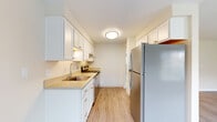 This single-floor residence offers a thoughtfully organized layout encompassing a combined kitchen and living room, one bedroom, one full bathroom, and two closets. The living room is a bright, open space finished with light-toned luxury vinyl plank flooring and a textured ceiling. Natural light pours in through a large picture window and an adjacent sliding glass door, both overlooking a green outdoor setting. A wall-mounted heater provides efficient climate control.

The galley-style kitchen runs parallel to the living area and features white shaker cabinetry extending to the ceiling, light stone countertops, a stainless steel sink, stainless steel refrigerator, and additional appliances including a range and microwave. Light wood-look flooring carries through the kitchen, contributing to a cohesive aesthetic.

The bedroom is a well-sized, quiet retreat carpeted in neutral tones and illuminated by a window with outdoor views. Two closets provide organized storage throughout the unit. The full bathroom includes a full-size soaking tub with an integrated surround, chrome fixtures, and a clean, functional design with warm neutral tones throughout.