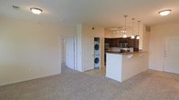 This single-floor residence showcases an open-concept layout that connects the living area and kitchen in one cohesive space. The living area is finished with plush wall-to-wall carpeting in a neutral tone, creating a warm and comfortable foundation throughout. A hallway visible from the main space leads to additional rooms, offering a practical and well-organized floor plan.

The kitchen is a standout feature, boasting rich dark-wood cabinetry that contrasts with a white peninsula island topped with a warm-toned countertop. The peninsula provides seating space, making it ideal for casual dining or entertaining. Stainless steel appliances, including a microwave and range, complement the kitchen's design. Overhead, a combination of pendant lights and a track lighting fixture provides focused illumination across the cooking and prep areas.

A notable convenience is the in-unit laundry closet, which houses a stacked washer and dryer tucked neatly between the kitchen and hallway. The neutral color palette throughout—cream walls, light ceilings, and toned carpeting—contributes to a clean, cohesive ambiance that feels both functional and inviting.