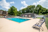 Located at 111 Whitsett Rd in Nashville, Tennessee, this outdoor pool facility offers a well-appointed recreational space designed for relaxation and leisure. The centerpiece is a generously sized swimming pool with clear blue water, accessible via pool steps and framed by a wide concrete sun deck that provides ample room for lounging and socializing. The deck is furnished with multiple table-and-chair sets in teal and navy, offering convenient spots for poolside gatherings, along with additional reclining lounge chairs positioned to take advantage of the open sky and sunlight. A covered pergola or shade structure is also present on the deck, providing a shaded retreat on sunny days. The area is enclosed by a chain-link security fence, with lush, mature trees lining the perimeter and creating a natural green backdrop that lends a sense of privacy and tranquility. A utility shed is located on the premises, and a notice board near the entry door provides informational postings for guests. The surrounding landscape features well-maintained hedgerows and open green lawn areas visible beyond the fencing. The overall setting is open-air, sun-filled, and thoughtfully equipped to serve as a comfortable community outdoor amenity in Nashville.