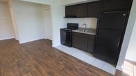 This single-floor space showcases an open layout where the kitchen transitions seamlessly into the living area. The kitchen is equipped with dark espresso-toned cabinetry that provides ample upper and lower storage. Black appliances, including a gas range and a full-sized refrigerator, complement the dark cabinetry for a cohesive look. A stainless steel sink is positioned beneath the upper cabinets, and a range hood is installed above the stove for ventilation. The kitchen features white tile flooring, which contrasts neatly with the warm, wood-style laminate flooring that extends throughout the adjacent living area. The transition between the two flooring materials creates a clear visual distinction between spaces. Light-toned walls throughout contribute to a bright and neutral ambiance. An arched doorway visible in the background leads to an additional room or hallway, hinting at further functional spaces within the unit. The overall design is practical and straightforward, with a focus on functionality and ease of movement between the kitchen and living areas.