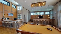 This well-appointed single-floor communal amenity space is designed to offer a dynamic and comfortable environment for socializing and recreation. The open-concept layout flows seamlessly between distinct activity zones, all unified by warm wood flooring, earthy tones, and an abundance of natural light streaming through an impressive array of clerestory and full-height windows that frame views of lush palm trees and greenery.

The game room is a standout feature, anchored by a full-size billiards table beneath a striking industrial-style pendant chandelier with amber glass shades. An elevated bar counter with backless wood-and-metal stools wraps around the perimeter, offering casual seating throughout the space.

Adjacent to the game area, a comfortable lounge provides a relaxed gathering spot furnished with plush sofas, accent chairs, and a rustic coffee table, all arranged around a large wall-mounted flat-screen television with built-in speakers — ideal for watching events or gaming together.

A compact but functional kitchenette serves the space with a stainless steel sink, generous countertop workspace, a coffee machine, and snack storage, making it well-suited for extended gatherings. A cozy bistro nook with a round high-top table and sculptural stools provides an intimate spot for small group conversation, completing this thoughtfully designed communal retreat.