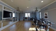 Located at 722 Island Park Drive on Daniel Island, SC, this single-floor fitness center offers a thoughtfully designed workout space that balances functionality with an inviting atmosphere. The room features warm hardwood flooring throughout, providing a clean and durable surface suited for a variety of exercise activities. Soft blue-gray walls lend a calm, focused ambiance to the space, while crown molding and white trim add a refined architectural touch.

Natural light filters in through multiple windows, brightening the interior and creating an open, energizing environment. Recessed ceiling lights and several industrial-style ceiling fans ensure the space remains well-lit and comfortable during workouts. A full wall of mirrors along one side visually expands the room and provides functional feedback for exercise form.

The space is well-stocked with equipment, including a row of stationary cycling bikes positioned toward the center, along with a well-organized selection of kettlebells, medicine balls, and free weights neatly arranged on racks along the wall. The generous floor plan allows ample room for movement between equipment, making it suitable for both group and individual fitness sessions.