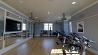 Located at 722 Island Park Drive on Daniel Island, SC, this single-floor fitness center offers a thoughtfully designed workout space that balances functionality with an inviting atmosphere. The room features warm hardwood flooring throughout, providing a clean and durable surface suited for a variety of exercise activities. Soft blue-gray walls lend a calm, focused ambiance to the space, while crown molding and white trim add a refined architectural touch.

Natural light filters in through multiple windows, brightening the interior and creating an open, energizing environment. Recessed ceiling lights and several industrial-style ceiling fans ensure the space remains well-lit and comfortable during workouts. A full wall of mirrors along one side visually expands the room and provides functional feedback for exercise form.

The space is well-stocked with equipment, including a row of stationary cycling bikes positioned toward the center, along with a well-organized selection of kettlebells, medicine balls, and free weights neatly arranged on racks along the wall. The generous floor plan allows ample room for movement between equipment, making it suitable for both group and individual fitness sessions.
