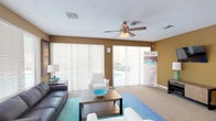 This single-floor space centers around a generously sized living area designed for comfort and functionality. The room is finished with wall-to-wall carpeting in a neutral tone, complemented by warm tan-painted walls that create an inviting atmosphere. A textured ceiling with a ceiling fan and light fixture provides both practical airflow and ambient lighting throughout the space.

Natural light is a defining feature, with large sliding glass doors dressed in vertical blinds that open to an outdoor pool area, offering pleasant views and easy access to the exterior. Additional windows fitted with horizontal blinds further brighten the interior. The living area accommodates a substantial seating arrangement, including a dark leather sofa, white accent chairs, and a rectangular coffee table centered on a vibrant teal area rug.

A wall-mounted flat-screen television sits above a low-profile media console on one side of the room, while decorative framed artwork adds visual interest to the walls. A secondary seating nook near the entrance features a side table with a lamp and additional accent chair. Tile flooring is visible near the entry area, transitioning into carpet within the main living space, offering a clear delineation between zones. The overall layout is open, practical, and well-lit.