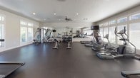 This single-floor fitness center is designed to provide a comprehensive workout experience in a bright, functional environment. The main exercise area is spacious and open, featuring durable dark rubber flooring that spans the entire space, providing a safe and comfortable surface for a wide range of activities.

Natural light floods the room through expansive floor-to-ceiling windows fitted with adjustable blinds, offering both illumination and outdoor views. A ceiling fan and recessed lighting ensure year-round comfort and visibility throughout the facility.

The cardio zone is well-appointed with multiple treadmills, elliptical machines, and a stationary bike, catering to various aerobic training preferences. A large wall-mounted mirror along one side of the room amplifies the sense of space and allows users to monitor their form during workouts. The strength training area includes a multi-station cable machine and a full rack of dumbbells, accommodating a variety of resistance exercises.

Along one wall, a practical utility area features dual stainless steel water fountains at varying heights, a wall-mounted paper towel dispenser, a storage cubby unit with bench seating for personal belongings, and coat hooks. A mounted wall clock and flat-screen TV add functional and entertainment value to the space. A separate restroom is accessible directly from the main fitness floor, enhancing the overall convenience of the facility.