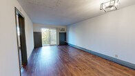 This well-organized one-bedroom, one-bathroom unit delivers a practical and comfortable single-floor layout across a compact footprint. The living room is an open, light-filled space featuring rich hardwood-style flooring, a textured ceiling, a modern geometric light fixture, and baseboard heating. A sliding glass door leads to a private balcony overlooking mature trees, while a wall-mounted air conditioning unit provides additional climate control. The kitchen is efficiently arranged in a galley-style configuration, featuring warm wood cabinetry, dark granite-look countertops, a double basin sink, and white appliances including a refrigerator, electric range, and dishwasher. The bedroom offers plush wall-to-wall carpeting, a ceiling fan with light, a dedicated closet with sliding gray panel doors and built-in shelving, and a window that brings in natural light. The bathroom is clean and bright, appointed with a full white-tiled tub and shower surround with a built-in niche and chrome fixtures. Throughout the unit, gray-painted accents on doors and trim add a consistent, modern touch that ties the overall aesthetic together.