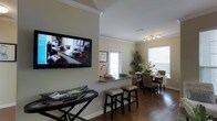 This single-floor residence showcases a thoughtfully designed open-concept layout that integrates the living, kitchen, and dining areas into one cohesive, functional space. Crown molding throughout adds a refined architectural detail, lending an air of elegance to the interior.

The living area features a wall-mounted flat-screen television and is anchored by a console table, creating a welcoming entertainment space. It flows naturally into a kitchen pass-through with a granite-topped breakfast bar, complete with bar stools — ideal for casual dining or conversation. The kitchen sink sits at the bar, reinforcing the open connection between spaces.

The dining area is positioned near large windows dressed with horizontal blinds, allowing abundant natural light to fill the room. A woven rattan dining set with seating for several guests adds a warm, organic texture to the space. A tall indoor plant in the corner contributes a fresh, natural element to the overall ambiance.

Rich hardwood flooring runs throughout, tying all areas together with a warm and consistent tone. Track lighting above the kitchen area provides focused illumination, while the neutral wall palette keeps the space feeling bright and airy. A glass door at the rear offers additional natural light and hints at outdoor access.