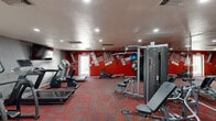 Located at 2601 East McKellips Road in Mesa, Arizona, this well-equipped fitness center offers a comprehensive workout environment within a single, open floor plan. The space is defined by its bold, energetic aesthetic — a striking red accent wall dominates one end of the room, covered in motivational typography including phrases like "Do It," "Dedication," and "Workout," creating an inspiring atmosphere for exercise.

The cardio section features multiple treadmills, an elliptical trainer, a stationary bike, and a rowing machine, all arranged to allow comfortable spacing between users. The strength training area includes a multi-station cable machine, a leg press, adjustable weight benches, and a medicine ball rack, providing a well-rounded selection of resistance equipment. A wall-mounted flat-screen television is positioned for visibility from the cardio machines.

Full-length mirrors line one wall, amplifying the sense of space and allowing users to monitor their form. The floor is covered in a durable, patterned carpet in grey and red tones that complements the room's color scheme. Recessed lighting provides even illumination throughout. A water dispenser station is available for hydration. The room is accessed through elegant French doors and includes a separate interior door, ensuring a clean, self-contained environment for focused fitness activity.