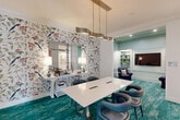 Located at 20013 Century Blvd in Germantown, MD, this sophisticated dining room offers a refined and visually distinctive interior design. The space is anchored by a sleek white rectangular table surrounded by contemporary navy blue upholstered chairs, positioned beneath a brushed gold linear pendant light that adds warmth and a modern elegance to the room. Underfoot, a vibrant teal distressed-style area rug ties the color palette together and adds a bold design statement throughout the space. A standout feature is the botanical-themed wallpaper adorning one wall, showcasing an intricate pattern of tropical birds, bamboo, and florals that lends a lively, artistic energy to the room. Two framed mirrors are mounted above a white console table along this wall, amplifying light and depth. The adjacent alcove area is finished in soft mint tones and features built-in shelving, a wall-mounted flat-screen television, and a plush navy accent chair — creating a comfortable secondary seating nook. Crown molding and wainscoting details throughout lend the space a polished, architectural character. Colorful bird-themed artwork punctuates the walls, and access to a bright, welcoming entry corridor with wood-framed glass doors and decorative tile flooring is visible from within the room.