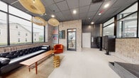 Located at 329 Totowa Rd in Totowa, New Jersey, this single-floor lobby space masterfully blends industrial character with contemporary design. Exposed light brick walls serve as a defining architectural element throughout, contrasted by a dark drop ceiling with recessed lighting and statement woven pendant fixtures that add warmth and visual texture overhead.

The entrance is anchored by floor-to-ceiling black-framed glass doors and expansive windows that flood the space with natural light and offer open views of the surrounding streetscape and greenery. Large-format white tile flooring transitions seamlessly across the main areas, while a raised platform with a dark stone surface adds architectural dimension near the entry.

A welcoming lounge area is thoughtfully furnished with a tufted black leather sofa, rich orange velvet armchairs, a mid-century wood coffee table, and organic wood accent stools — all arranged atop a woven area rug. Bold framed artwork punctuates the brick walls, adding curated character to the space.

Toward the rear, a bank of individual mailboxes is set against a striking dark marbled accent wall, accompanied by a glass-paneled door providing access to the stairwell. Floating black shelves mounted on brick offer functional display space near the entrance. The overall ambiance is polished yet inviting, combining urban-industrial aesthetics with modern, residential-quality comfort.