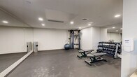 This state-of-the-art fitness facility, located at 500 Asher Rd in Kelowna, BC, offers a compact yet comprehensive workout space within its 41 square meters. The room boasts a sleek, modern design with white textured walls and smooth concrete flooring, creating a clean and minimalist aesthetic. Bright, recessed LED lighting illuminates the space evenly, enhancing visibility and energy.

The gym is well-equipped with a range of exercise machines, including multiple treadmills, weight training equipment, and cardio machines. A large wall-mounted mirror spans one side of the room, allowing users to monitor their form during workouts. The space also includes functional training areas with stability balls and free weights.

Safety features are evident, with a visible fire alarm system and strategically placed waste receptacles for convenience. Large windows provide natural light and a connection to the outdoors. The layout is thoughtfully designed to maximize the available space while ensuring ample room for movement between equipment. This fitness center combines functionality with modern design, offering an inviting environment for health and wellness activities.