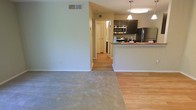 This single-floor space showcases an open-concept layout that seamlessly connects the living area to the kitchen, creating a cohesive and functional environment. The living area is finished with soft, neutral-toned carpet, providing a comfortable and understated foundation for the space. A clear transition to hardwood-style flooring marks the shift into the kitchen and hallway areas, adding visual interest and practicality.

The kitchen is well-appointed with dark espresso cabinetry that contrasts neatly against the light walls, and it is equipped with stainless steel appliances including a refrigerator, range, and over-the-range microwave. A peninsula with granite-style countertops serves as both a prep area and a casual dining or gathering spot, enhanced by pendant lighting overhead that adds warmth and definition to the space.

The walls throughout are painted in a soft, muted sage-green tone, contributing to a calm and neutral ambiance. A hallway visible from the main living area leads further into the unit, suggesting additional rooms beyond the open living and kitchen zone. Ceiling vents indicate a central HVAC system. Overall, the space presents a clean, functional, and well-maintained interior.