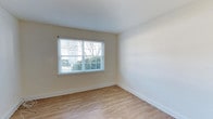 This single-story space offers a practical and well-organized layout with a clean, neutral aesthetic throughout. The kitchen is equipped with dark espresso-toned cabinetry that extends to the upper walls, providing ample storage, and features stainless steel appliances including a full-size refrigerator. A louvered bifold door adds a classic architectural detail near the kitchen entry, alongside a wall-mounted thermostat for climate control. Light hardwood-style flooring runs throughout, lending warmth and visual continuity across the space. The hallway connects the kitchen area to additional rooms, maintaining an open and navigable flow. A separate room, finished with the same light flooring and crisp white baseboards, is bathed in natural light from a large window fitted with horizontal blinds. The window frames an outdoor view of mature trees and greenery, bringing a sense of the outdoors inside. Walls throughout are painted in soft, neutral tones, complementing the light flooring and creating a bright, welcoming ambiance. The overall design is straightforward and functional, with consistent finishes that give the space a cohesive and move-in-ready feel.