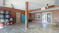 This single-floor space offers an open, functional layout designed for practical use. The interior is defined by striking deep burgundy-red walls that create a bold, energetic atmosphere throughout. Dark hardwood-style flooring runs across the open area, adding warmth and a sense of continuity to the space.

Structural support columns anchored with white base trim are positioned throughout, contributing to the architectural character of the room. Ceiling fans are installed to ensure comfortable air circulation, and recessed lighting supplements the natural light that filters in through a glass-panel exterior door, which offers views to the outdoors.

A service or reception window framed in a lighter trim is set into the wall, suggesting a counter or office function within the space. Framed artwork displayed near the window adds a welcoming touch. Utility elements such as an FACP (Fire Alarm Control Panel) door and visible equipment storage indicate that the space is equipped with essential building systems.

The overall layout is open and versatile, with ample floor space suitable for a variety of uses such as community gatherings, events, or organizational activities. The combination of functional design, bold color choices, and practical features makes this an adaptable and well-equipped single-story space.