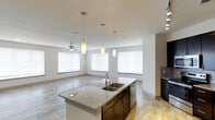 Located in Frisco, TX, this single-floor residence showcases a thoughtfully designed open-concept layout that seamlessly connects the kitchen and living areas. The kitchen is a standout feature, centered around a generous granite-topped island with a double basin sink and elegant pendant lighting that hangs above, creating both a functional workspace and a stylish focal point. Dark cabinetry provides a rich contrast against the light granite countertops, while stainless steel appliances and a tasteful tile backsplash add a polished, contemporary finish. The flooring throughout transitions between warm wood-look tile and lighter stone-toned tile, lending visual interest and continuity across the open space. The living area benefits from an abundance of natural light streaming through multiple large windows dressed with clean white blinds. A ceiling fan overhead enhances comfort and airflow in the living space. The neutral color palette of soft whites and warm grays throughout creates a calm, inviting atmosphere that is both modern and versatile. The overall design reflects a commitment to open, functional living with quality finishes, making this Frisco residence a well-appointed and comfortable space.