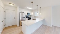 Located in Rehoboth Beach, Delaware, this one-bedroom, one-bathroom residence offers a thoughtfully designed single-floor layout with a clean, contemporary aesthetic. The open-concept living and kitchen area is bathed in natural light from multiple windows dressed with white blinds, creating an airy and inviting atmosphere. Light-toned hardwood-style flooring flows through the main living spaces, enhancing the sense of openness.

The kitchen is a standout feature, anchored by a large white quartz peninsula with an undermount sink and stylish pendant lighting overhead. White shaker-style cabinetry lines the walls, complemented by a mosaic tile backsplash and a full suite of stainless steel appliances, including a refrigerator, range, and microwave. The seamless connection between the kitchen and living area makes the space ideal for both everyday living and entertaining.

The bedroom is generously proportioned, finished with plush carpet, a ceiling fan, and abundant closet space provided by a set of double bi-fold doors. A wall-mounted HVAC unit ensures year-round comfort. The bathroom is clean and functional, featuring light wood-look flooring, a full tub-shower combination, and a vanity with ample storage. The overall design is bright, modern, and well-appointed throughout.