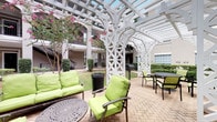 This single-floor space masterfully balances interior sophistication with inviting outdoor living. The interior dining area showcases a striking green marble round pedestal table paired with woven rattan chairs, all set atop a textured natural fiber rug over polished concrete floors. Warm recessed lighting illuminates the neutral cream-toned walls, which are adorned with a large-scale framed abstract artwork that adds an artistic focal point to the room. A rustic sliding barn door and a set of aged, character-rich double doors provide stylish transitions between spaces, hinting at additional rooms filled with curated wall art and décor visible beyond the thresholds. The interior exudes a curated, eclectic aesthetic that blends raw industrial elements with refined furnishings. Outside, a generously proportioned courtyard features an elegant white pergola with intricate lattice detailing, creating a sheltered and airy outdoor retreat. Vibrant green-cushioned seating arrangements invite relaxation, while additional café-style tables and chairs offer flexible gathering options. Mature flowering trees and manicured greenery frame the space, enhancing the sense of a private, lush oasis. Together, the interior and exterior areas create a cohesive, design-forward environment ideal for both everyday living and entertaining.