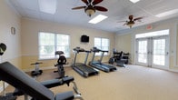 This single-floor fitness center is thoughtfully organized into two interconnected areas divided by elegant French doors. The primary cardio room houses multiple treadmills, a stationary recumbent bike, and a wall-mounted television, making it ideal for sustained aerobic workouts. A full dumbbell rack provides convenient access to free weights, while double glass doors open to an outdoor area, allowing fresh air and additional natural light to fill the space.

The secondary exercise room offers a more versatile environment, featuring a adjustable workout bench, cable resistance stations, a speed bag, and an additional recumbent bike. This area also includes open floor space suitable for stretching or group activities. A bookshelf and seating area with chairs provide a practical transition zone between rooms.

Throughout both areas, neutral beige walls with white wainscoting create a clean, welcoming atmosphere. Textured carpet flooring offers comfort underfoot, while multiple windows ensure consistent natural light. Ceiling fans in each room maintain air circulation. The interconnected layout and diverse equipment selection make this fitness center well-suited for a broad range of exercise routines.