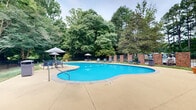 Located at 5425 Dana Dr in Raleigh, NC, this outdoor pool area offers a well-appointed recreational space surrounded by mature trees and natural greenery. The centerpiece is a generously sized freeform swimming pool with vivid turquoise water, featuring a curved, organic shape that adds visual interest to the space. A concrete pool deck wraps around the perimeter, providing ample room for lounging and socializing.

The deck is furnished with multiple chaise lounges and patio chairs, complemented by several shade umbrellas positioned throughout the space — ideal for relaxing out of the sun. A covered pergola or pavilion structure is also visible, offering additional shaded seating. The pool is enclosed by a wrought iron fence with brick accent walls, ensuring safety while maintaining an open, airy feel.

Adjacent to the pool is a brick utility or amenity building with a pitched roof, which appears to serve as a support facility for pool users. Tall pine trees and dense deciduous foliage frame the entire area, creating a private, park-like atmosphere. The lush natural surroundings provide a serene and shaded backdrop, enhancing the overall ambiance of this inviting outdoor space.
