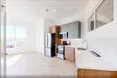 Located at 17 Avenue E in Bayonne, NJ, this thoughtfully designed one-bedroom, one-bathroom residence offers a clean, modern aesthetic throughout. Light oak-toned hardwood flooring and crisp white walls create a bright, cohesive atmosphere across the entire floor plan.

The open-concept kitchen and living area serves as the heart of the home. The kitchen is outfitted with stainless steel appliances — including a gas range, microwave, and refrigerator — complemented by warm wood cabinetry on the lower level and sleek matte gray upper cabinets. White quartz countertops and a glossy white tile backsplash complete the polished look, while a pass-through window adds an architectural detail that enhances the sense of openness.

The bedroom is generously proportioned, bathed in natural light from large floor-to-ceiling windows that frame outdoor views of the surrounding Bayonne neighborhood. A glass door leads directly to a private balcony, extending the living space outdoors. Two closets provide ample storage.

The bathroom features a full soaking tub with a glossy gray subway tile surround and contemporary fixtures. Additional conveniences include a dedicated laundry area and a utility room. The overall design is clean, minimal, and move-in ready.